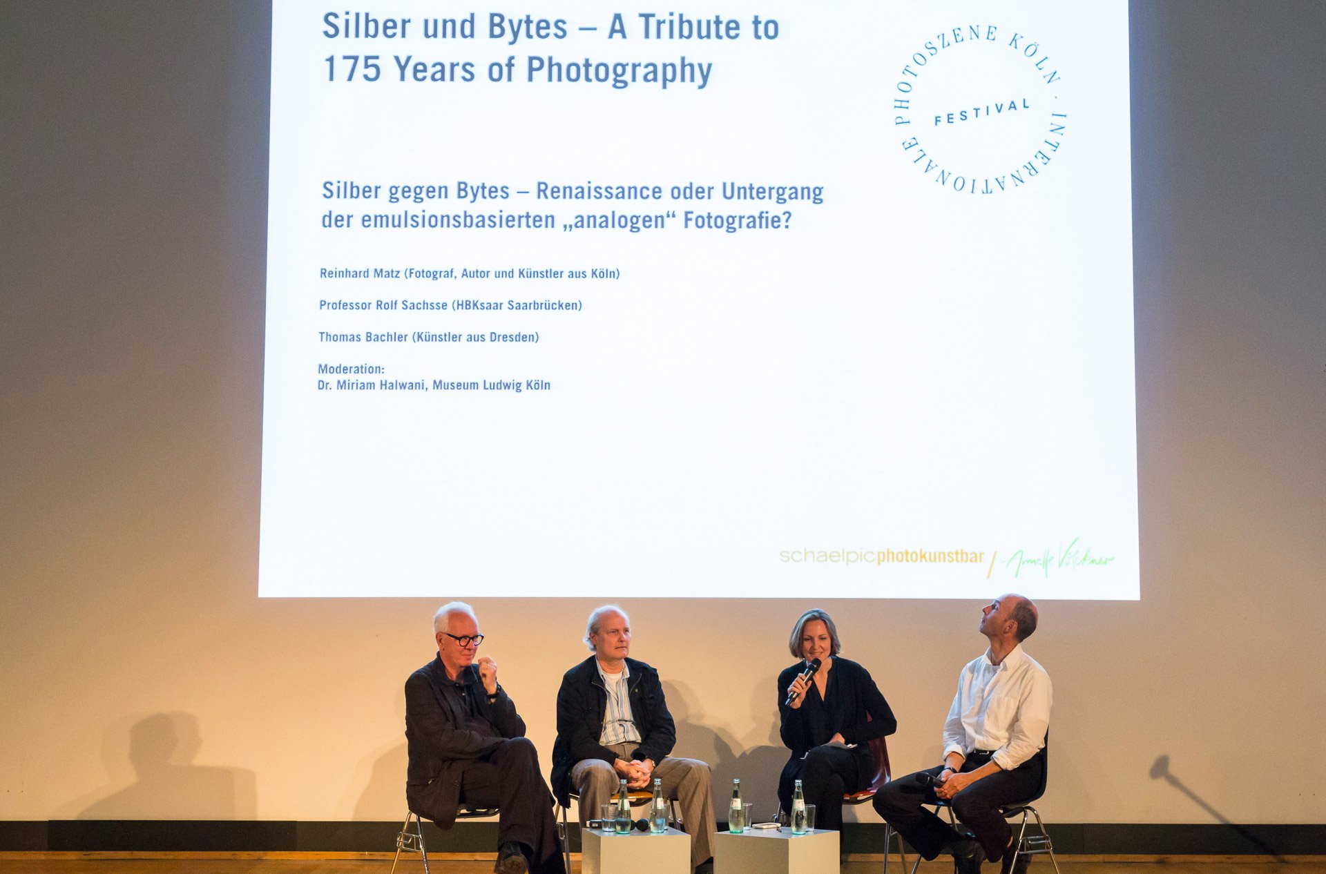 Silber und Bytes | Silber gegen Bytes; Podiumsdiskussion: Rolf Sachsse (HBKsaar Saarbrücken), Reinhard Matz (Fotograf, Autor und Künstler aus Köln), Miriam Halwani (Museum Ludwig), Thomas Bachler (Künstler aus Dresden) (v.li.). (Foto: Markus Bollen, 9/2014) Auf einem Podium sitzen vier Personen: Rolf Sachsse (HBKsaar Saarbrücken), Reinhard Matz (Fotograf, Autor und Künstler aus Köln), Miriam Halwani (Museum Ludwig), Thomas Bachler (Künstler aus Dresden) (v.li.). An die Wand hinter ihnen ist projiziert ›Silber und Bytes – 175 Years of Photography‹. (Foto: Markus Bollen, 9/2014)
