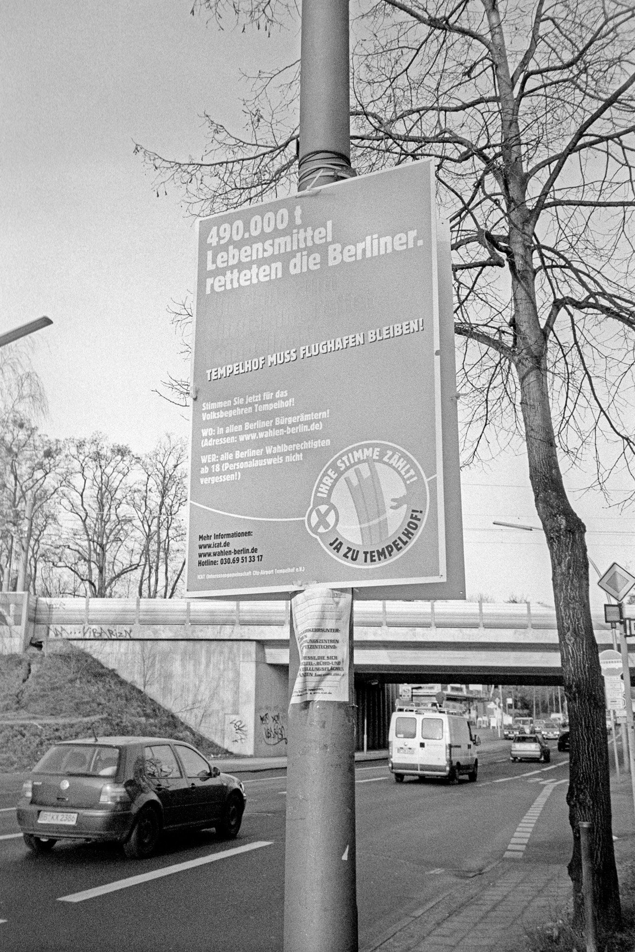 Schwarzweißbild: Plakat zum Volksbegehren an einem Pfahl in Berlin-Lankwitz, Leonorenstraße: ›Tempelhof muss Flughafen bleiben!‹ Im Hintergrund eine Eisenbahnbrücke. (Foto: Martin Frech, 1/2008)