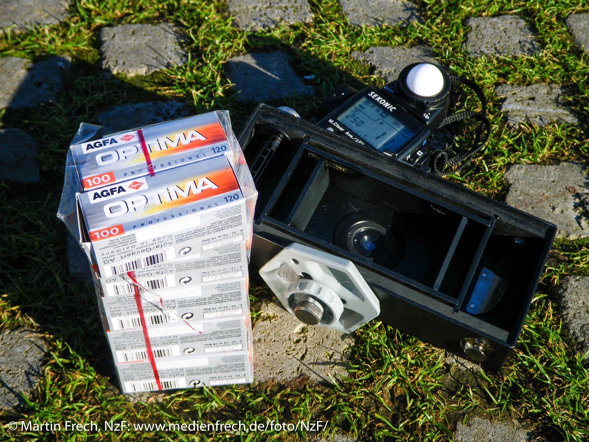 Farbbild: Ein angebrochener Zehnerpack Agfa Optima 100 liegt auf der Wiese vor dem Reichstagsgebäude in Berlin, daneben eine Lochkamera und ein Belichtungsmesser. (Foto: Martin Frech, 11/2008)