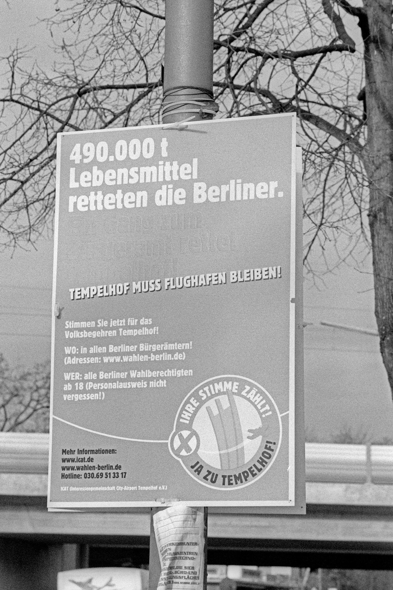 Schwarzweißbild: Plakat zum Volksbegehren an einem Pfahl in Berlin-Lankwitz, Leonorenstraße: ›Tempelhof muss Flughafen bleiben!‹ Im Hintergrund eine Eisenbahnbrücke. (Foto: Martin Frech, 1/2008)