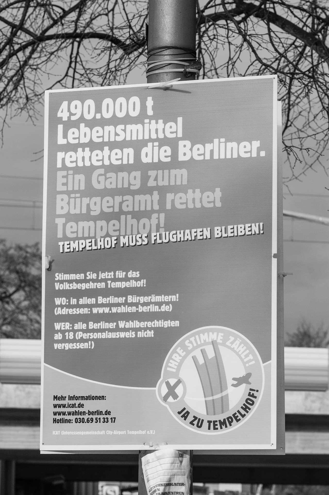 Schwarzweißbild: Plakat zum Volksbegehren an einem Pfahl in Berlin-Lankwitz, Leonorenstraße: ›Tempelhof muss Flughafen bleiben!‹ Im Hintergrund eine Eisenbahnbrücke. (Foto: Martin Frech, 1/2008)